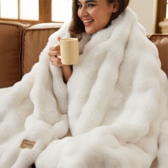 A woman wrapped in a white faux fur blanket holding a mug, setting a cozy scene.